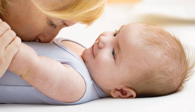 ©istock.com/simonkr Eine Frau mit blondem Haar küsst sanft die Nase eines lächelnden Babys, das auf dem Rücken liegt, beide auf einer weichen weißen Unterlage. Das Baby schaut die Frau an, die einen hellblauen ärmellosen Strampler trägt.